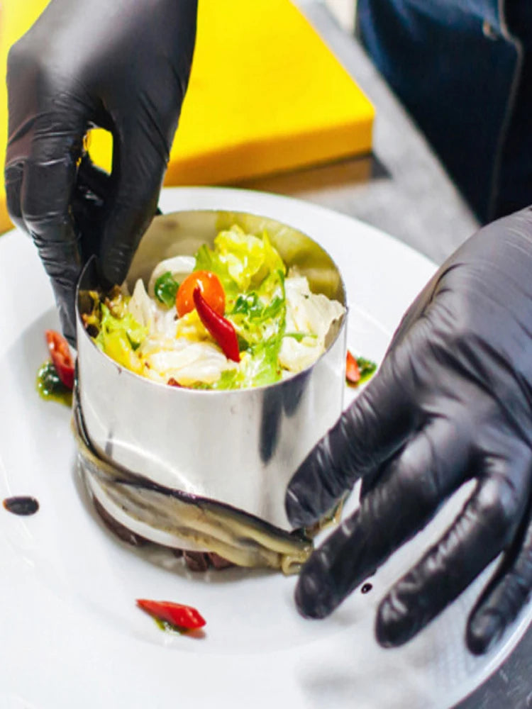 A hand wearing a black nitrile glove is holding a silver foil-wrapped dish containing a salad on a white plate.