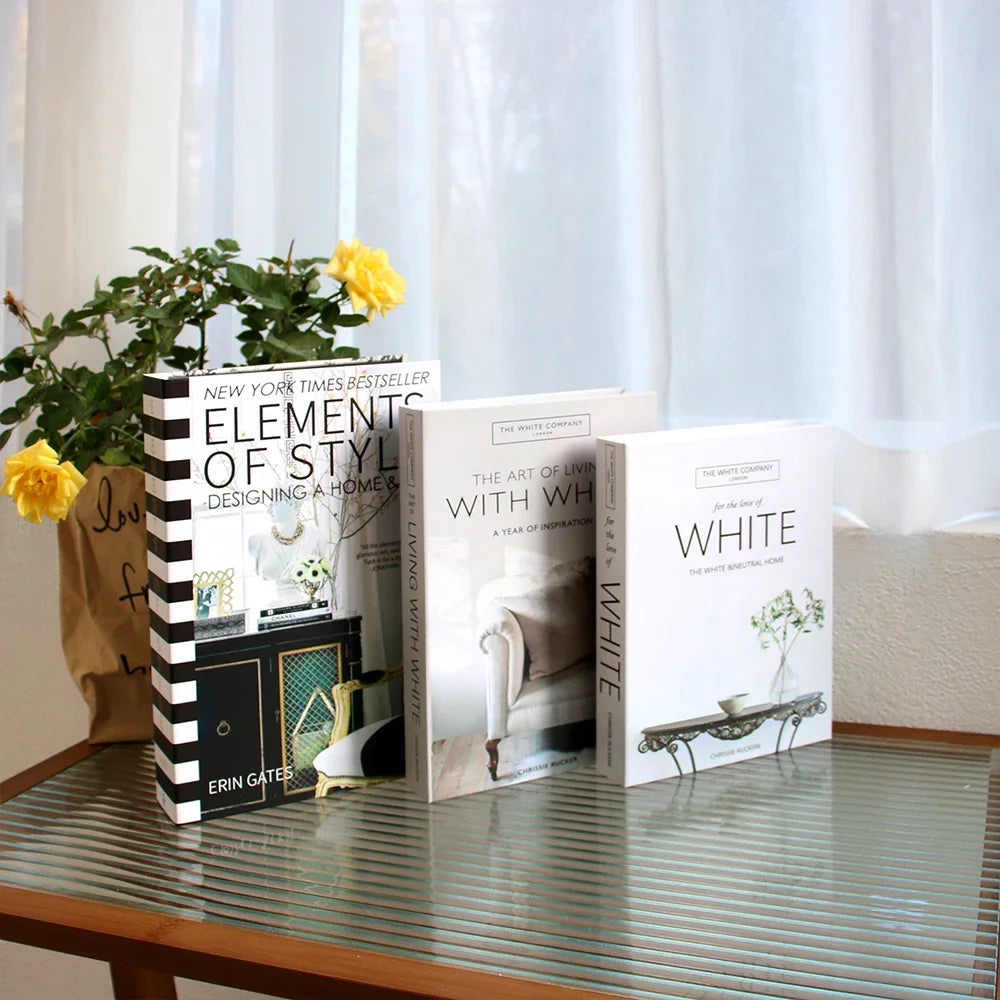 A stack of decorative fake books titled Elements of Style and The Art of Living with White set on a glass table.