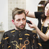 A woman is adjusting a black professional salon hair cutting cape on a man's neck.