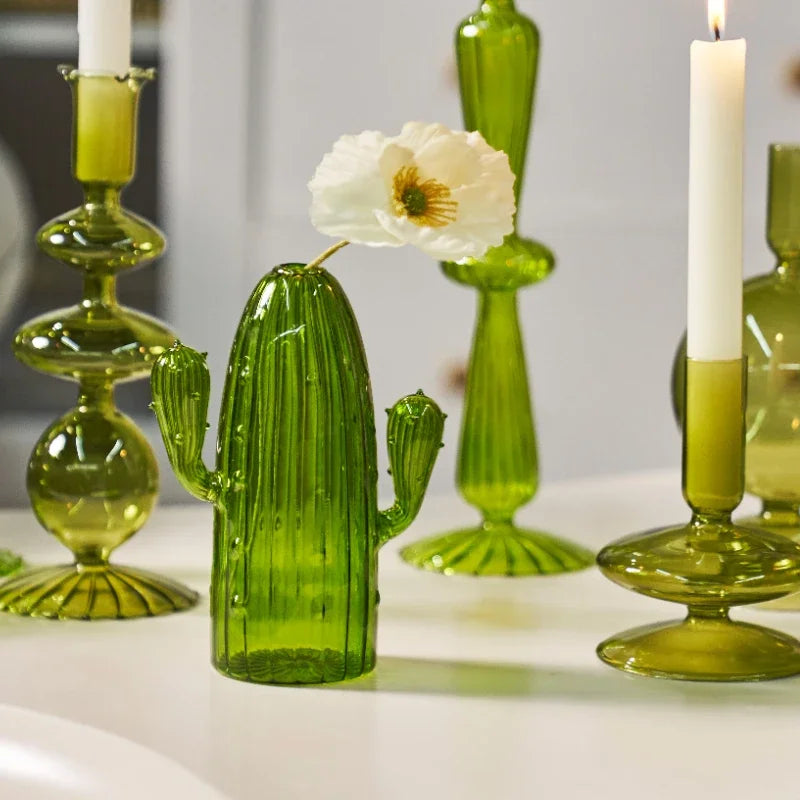 Un vase de cactus en verre moderne vert vif à côté de hauts chandeliers à bougies en verre et d'une fleur blanche.