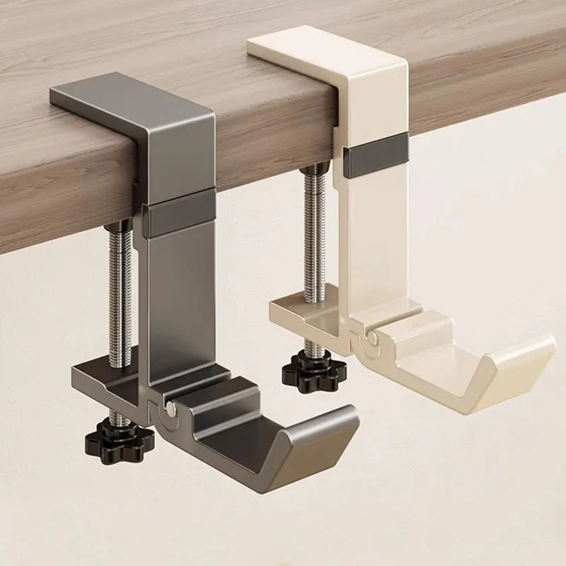 A close-up view of a metal and cream colored adjustable desk hook bag hanger mounted on a wooden desk surface.