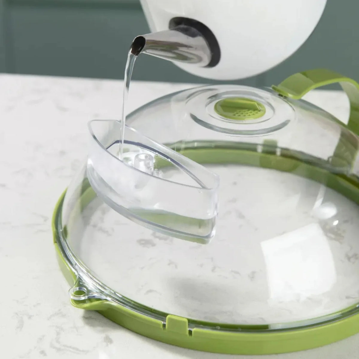 A clear microwave splatter cover with a water steamer is shown being used to pour liquid into a dish.