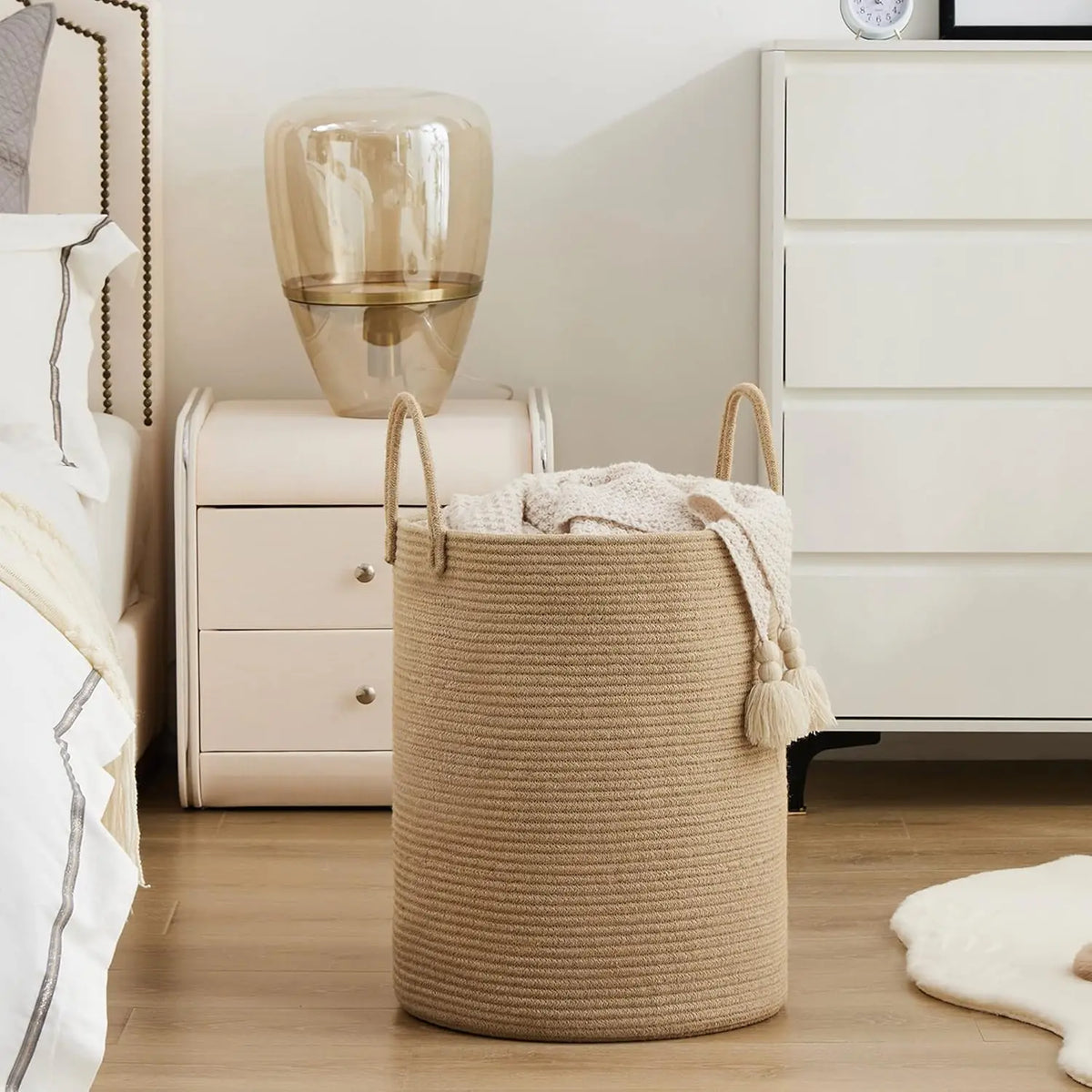 A large woven storage hamper with handles in a natural beige color sits on a wooden floor next to a white dresser and a glass
