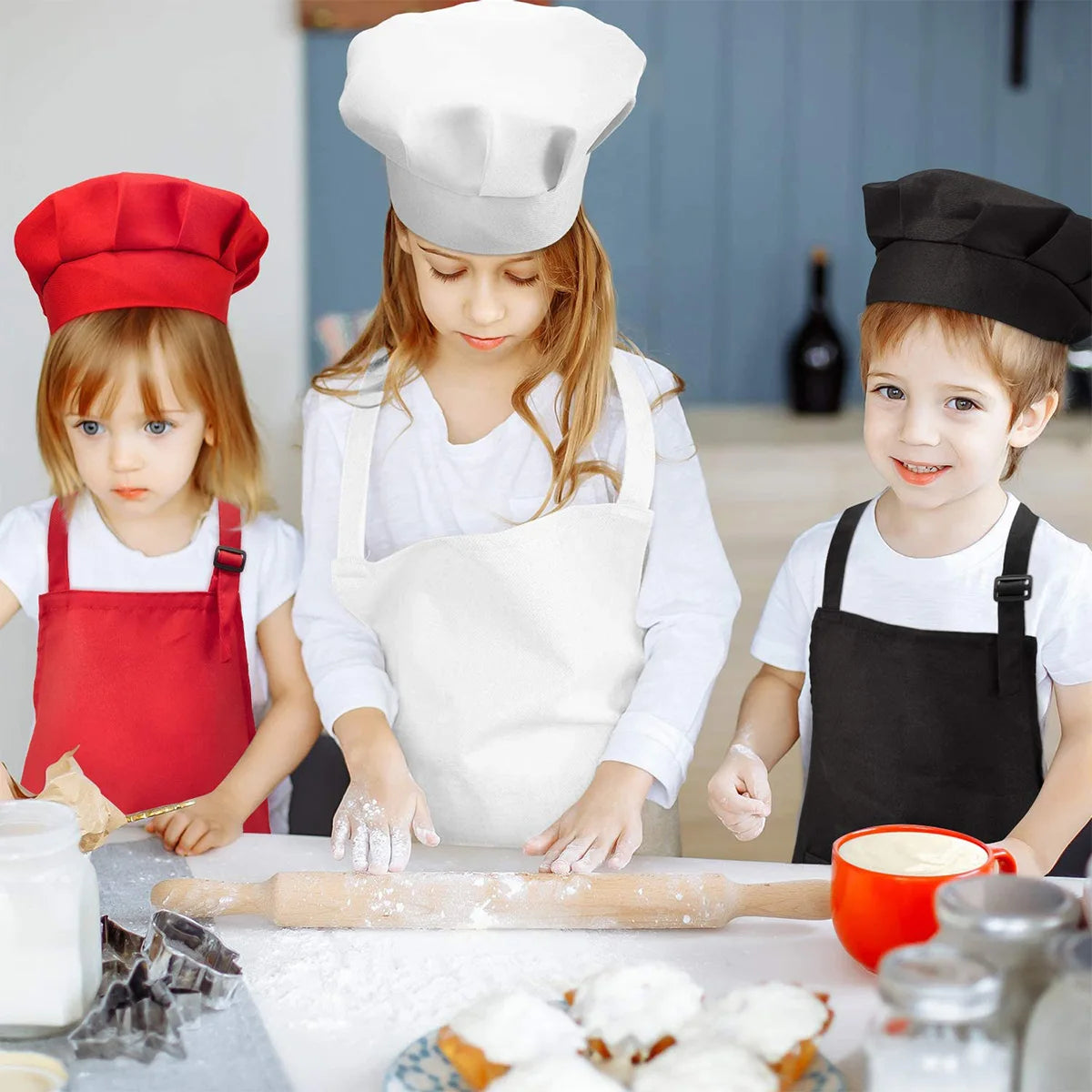 Two children are preparing food with a rolling pin and flour on a countertop wearing white aprons and chef hats.