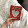 Un álbum de fotos rojo con ventana en forma de corazón y letras doradas, sostenido en una mano sobre ropa de cama blanca.
