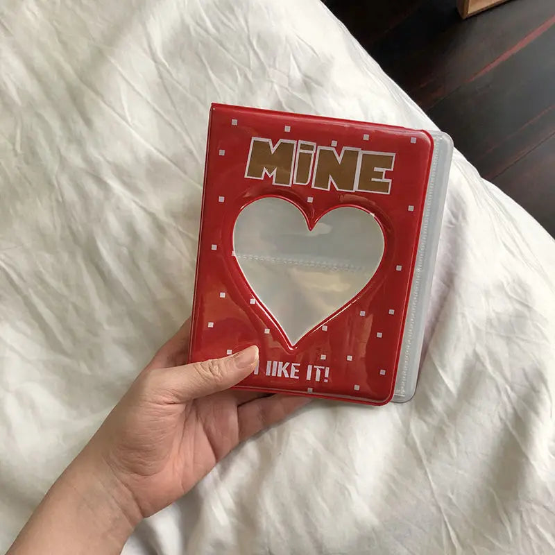 Un álbum de fotos rojo con ventana en forma de corazón y letras doradas, sostenido en una mano sobre ropa de cama blanca.