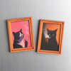 Zwei gerahmte Kunstdrucke mit schwarzen Katzen in rosa und orange Tönen.