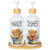 A white soap dispenser bottle with a wooden pump and a cow design labeled HANDS, sitting on a wooden tray.