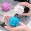 Three reusable laundry balls in pink, white, and blue are held in a person's hand against a gray fabric background.