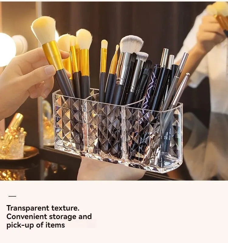 A hand holds a clear acrylic makeup brush holder with three compartments filled with various makeup brushes.