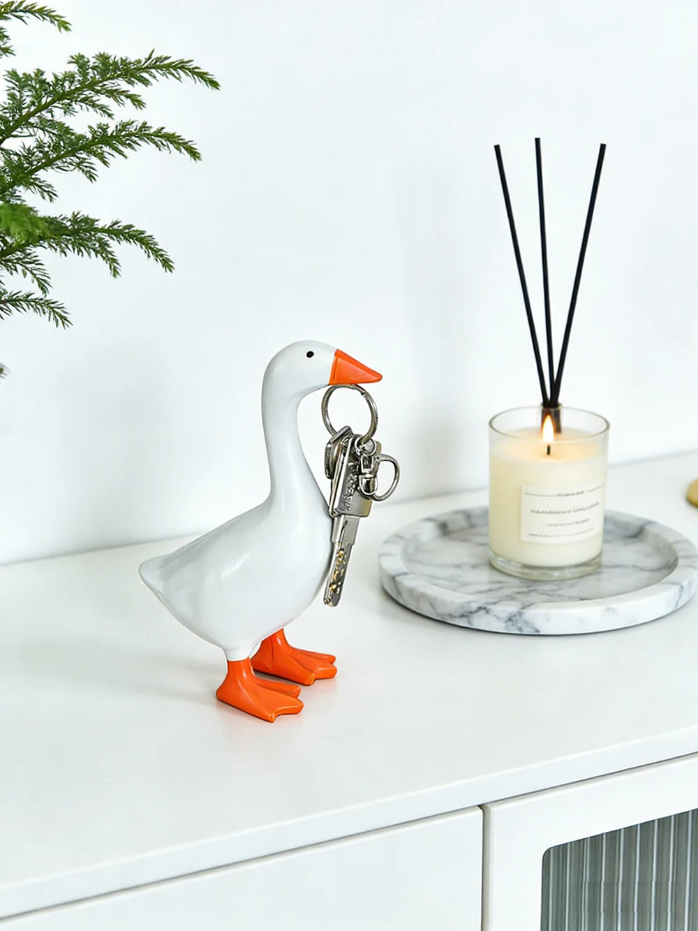 A white goose sculpture with an orange beak and legs stands next to a glass candle holder and a black key chain.