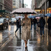 A woman in a beige trench coat holds a large black umbrella on a wet city street with other people using umbrellas.