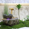 A miniature landscape featuring a moss-covered rock, a bamboo pipe, and a leafy plant on a white surface.