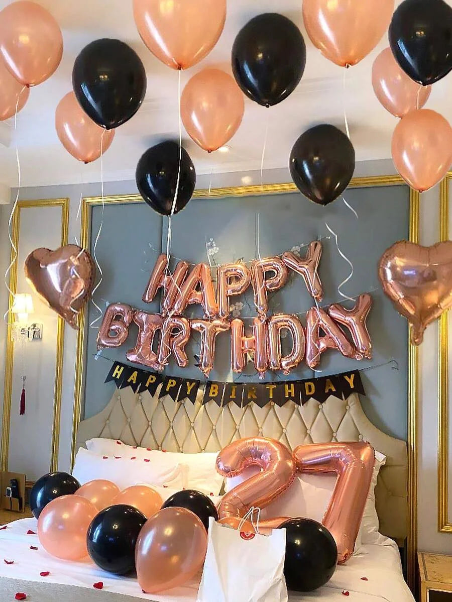 A celebration setup featuring rose gold and black foil balloons spelling out Happy Birthday, with a large number 7 on the bed