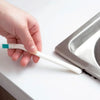 A hand holds a white silicone cleaning brush with a green connector, next to a metal bottle brush holder.
