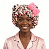A woman wearing a leopard print satin-lined sleep bonnet with pink ruffles and a pink pompom.