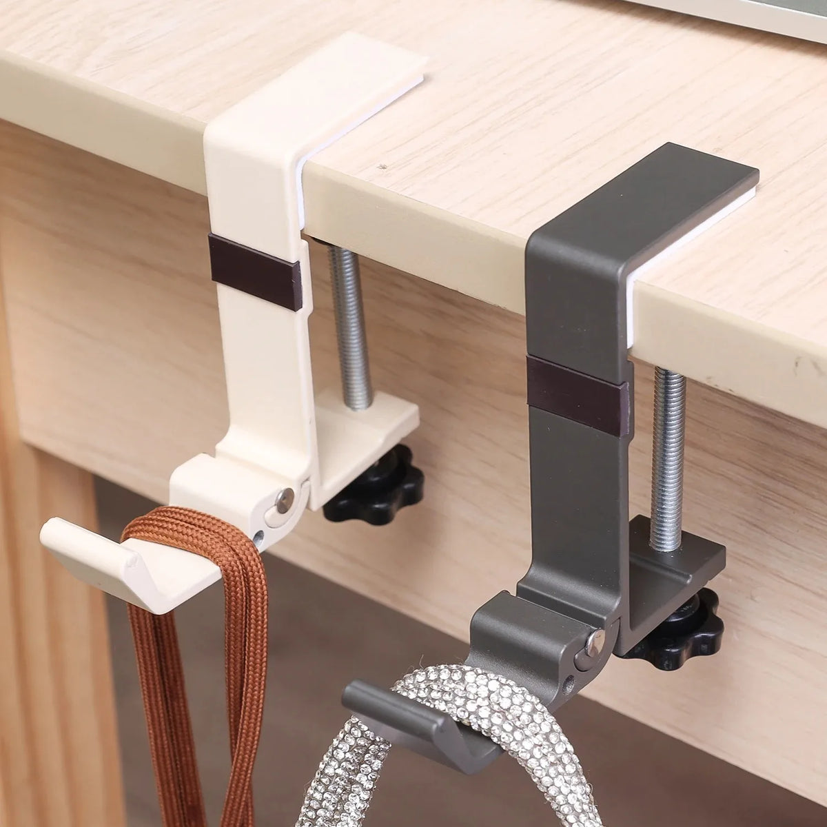 A close-up of an adjustable desk hook bag hanger with white and gray components mounted on a wooden desk surface.