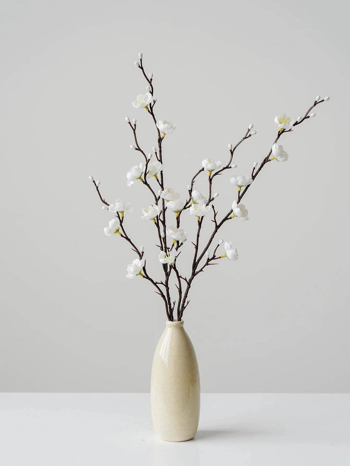 Eine schlanke Zweig künstlicher weißer Pflaumenblüten in einer hellen Keramikvase vor neutralem Hintergrund.