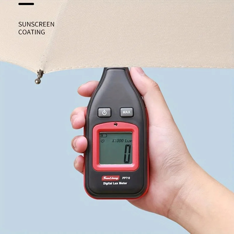 A hand holds a black digital lux meter device against a light beige umbrella canopy.