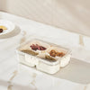 A clear plastic snack organizer tray with eight compartments filled with various dried snacks on a white marble countertop.