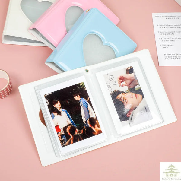 A white mini photo album with a clear cover is shown next to a pink and blue heart-shaped accessory.