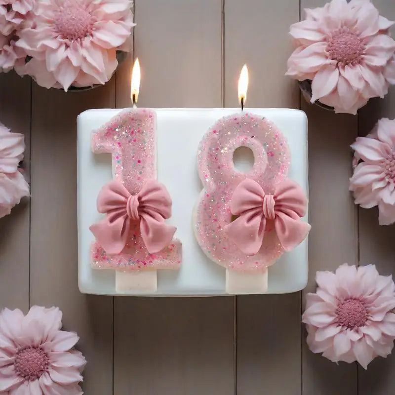 A white candle holder displaying glitter numbers 1 and 8, decorated with pink bows, set against a wooden background.