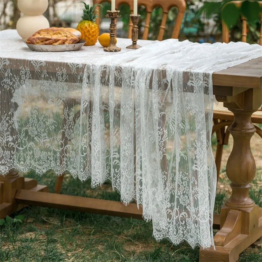 A table covered with a sheer white lace tablecloth set outdoors for a party.