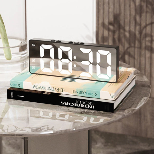 A digital LED mirror table clock displaying 08:30 sits on a glass table next to a stack of books.