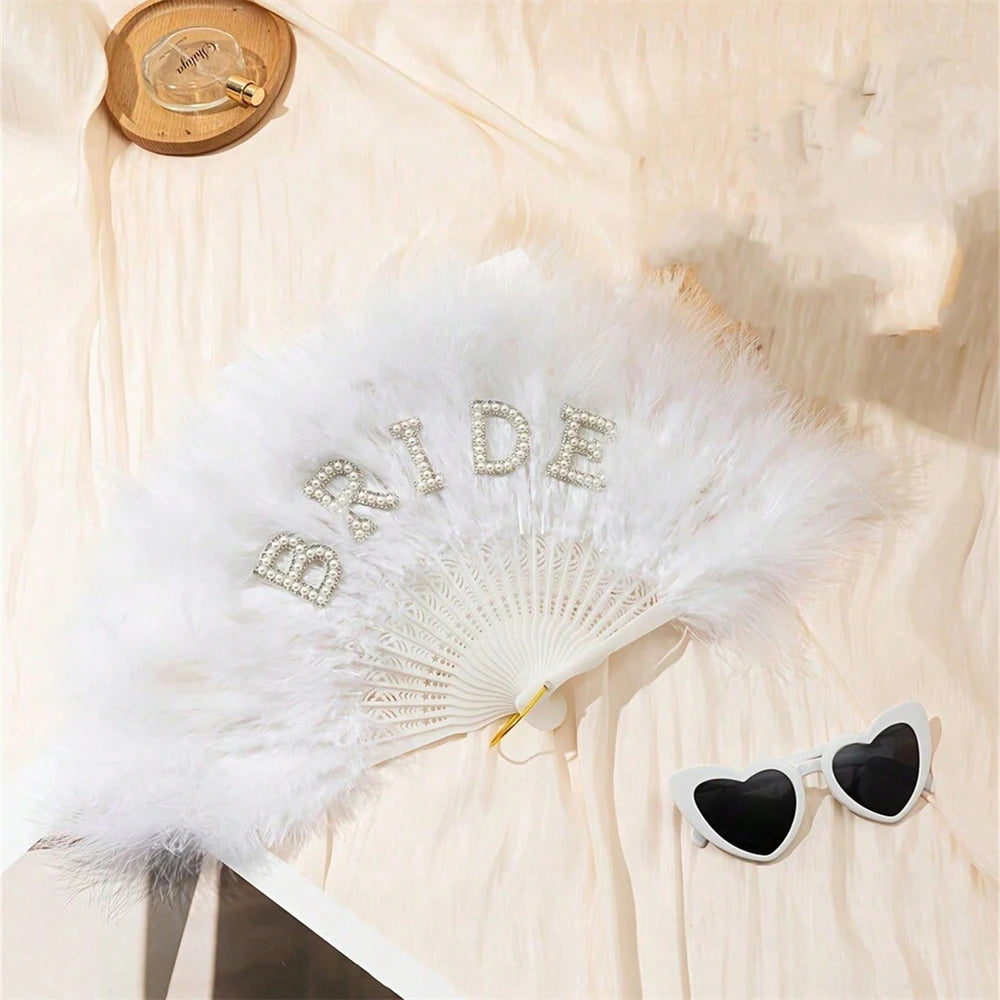 A white feather hand fan decorated with sparkling letters spelling bride on a light background.