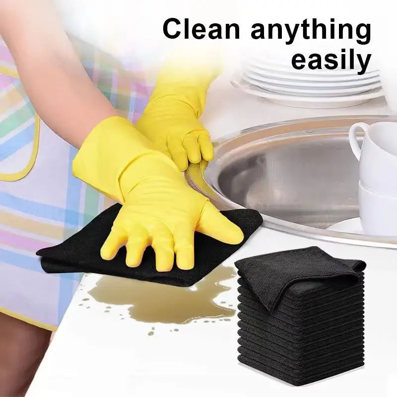 A hand wearing a yellow cleaning glove is wiping a stainless steel pan with a black microfiber cloth.
