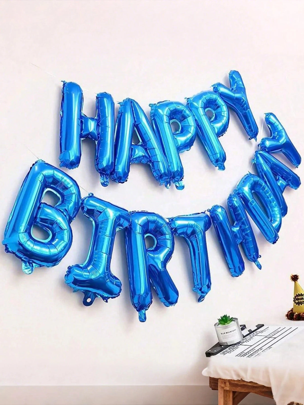 A silver and blue foil balloon banner spelling out Happy Birthday on a white wall.