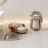 A clear plastic soap dish and container with a white bar of soap and a green soap dish resting on a white surface.