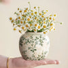 A hand holds a white ceramic bud vase filled with yellow and white daisies.