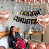 A woman is sitting on a bed surrounded by rose gold balloons and a banner that reads Happy Birthday.