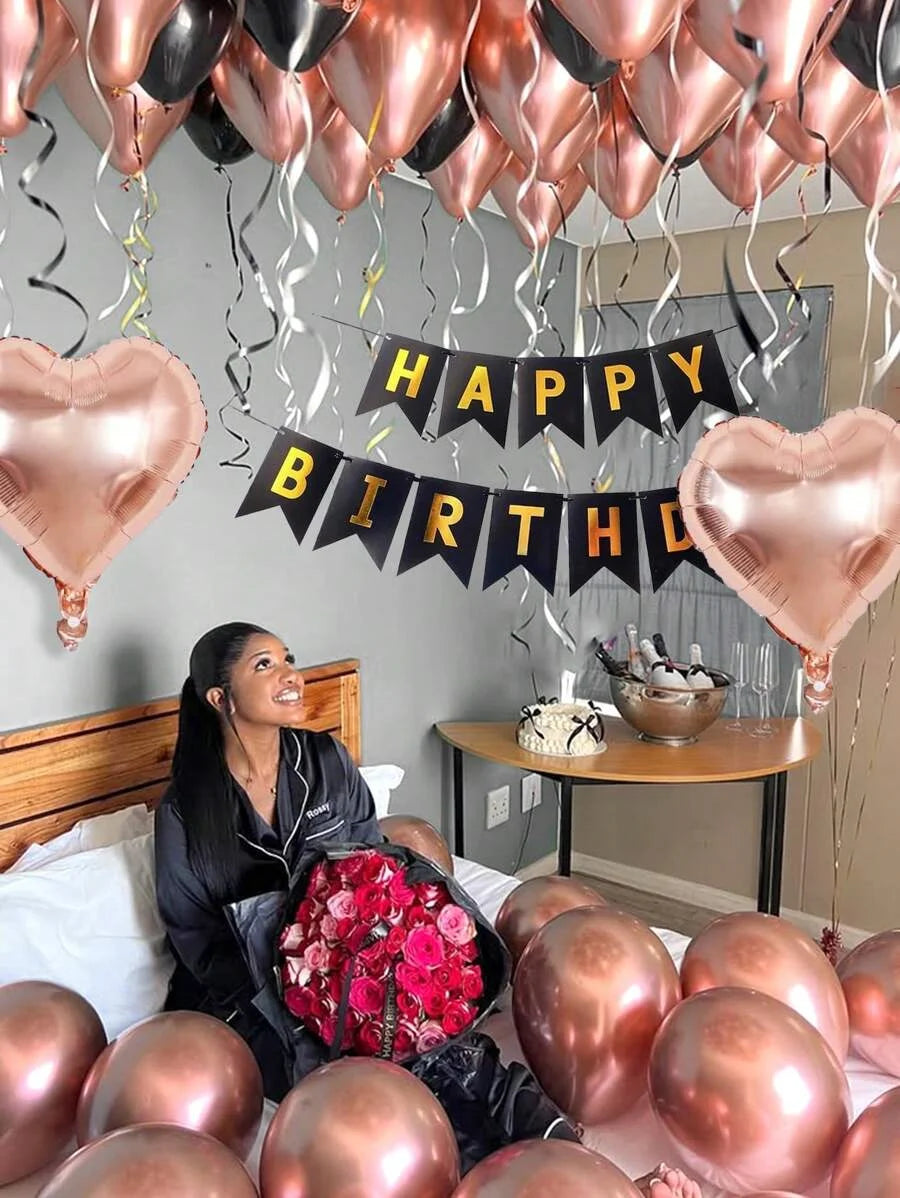 A woman is sitting on a bed surrounded by rose gold balloons and a banner that reads Happy Birthday.