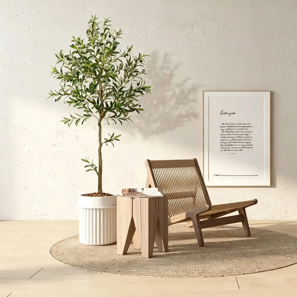 An artificial olive tree in a white pot stands next to a wooden chair and framed text on a light-colored wall.