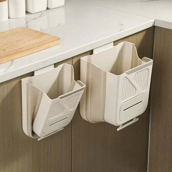 A beige foldable wall-mounted trash can is shown on a kitchen countertop next to a wooden cutting board.