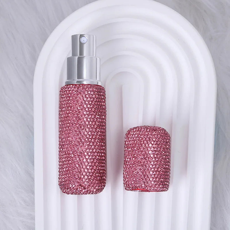 A glass perfume spray bottle with a silver pump and a textured pink glitter body is displayed on a white, ribbed stand.