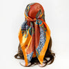 A women's square paisley print scarf tied around a dark hair, featuring orange, blue, and gold patterns.