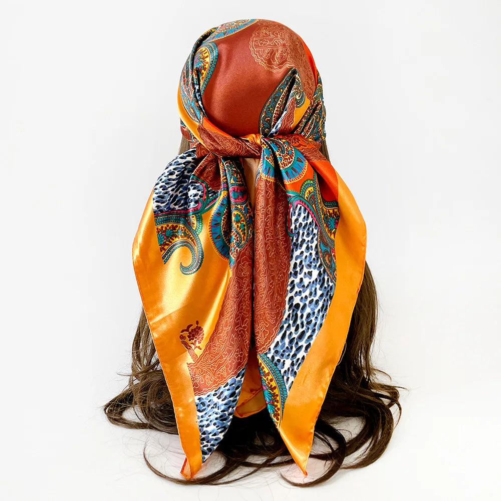 A women's square paisley print scarf tied around a dark hair, featuring orange, blue, and gold patterns.