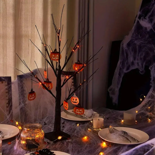 Dunkler Birkenbaum mit orangefarbenen LED-Lichtern und Halloween-Dekorationen auf einem Tisch.