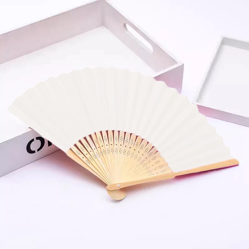 Un éventail pliable beige clair à la main, fait de bambou et de papier, partiellement ouvert sur fond blanc.