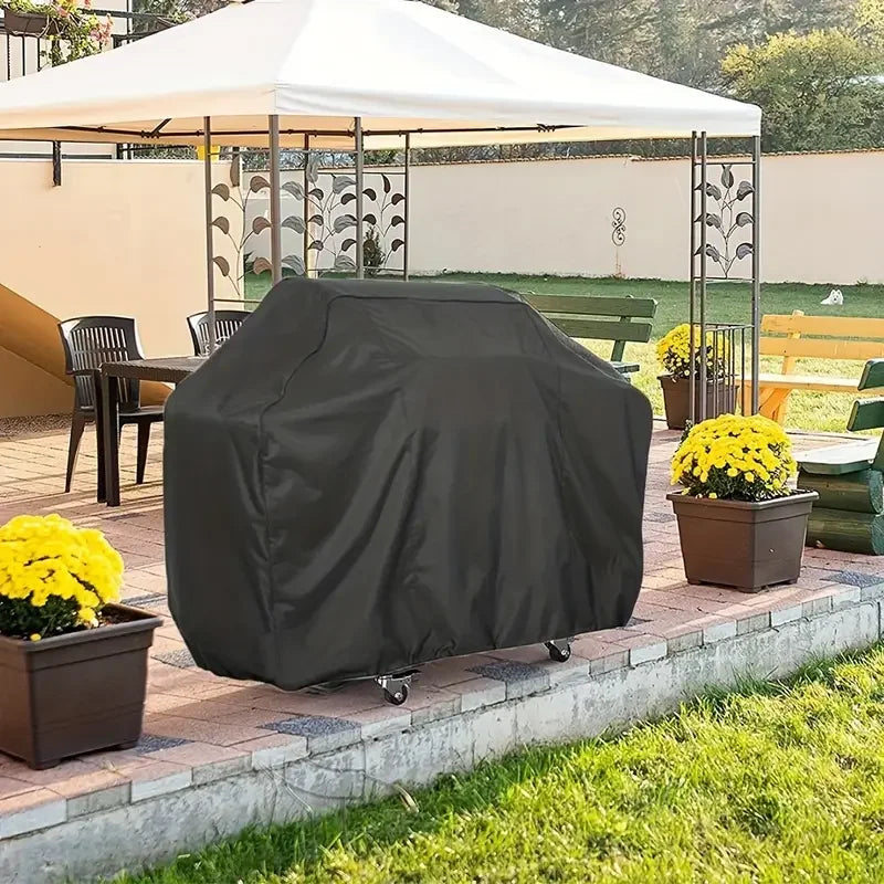 A dark gray heavy-duty grill cover is placed on a brick patio next to potted yellow flowers.