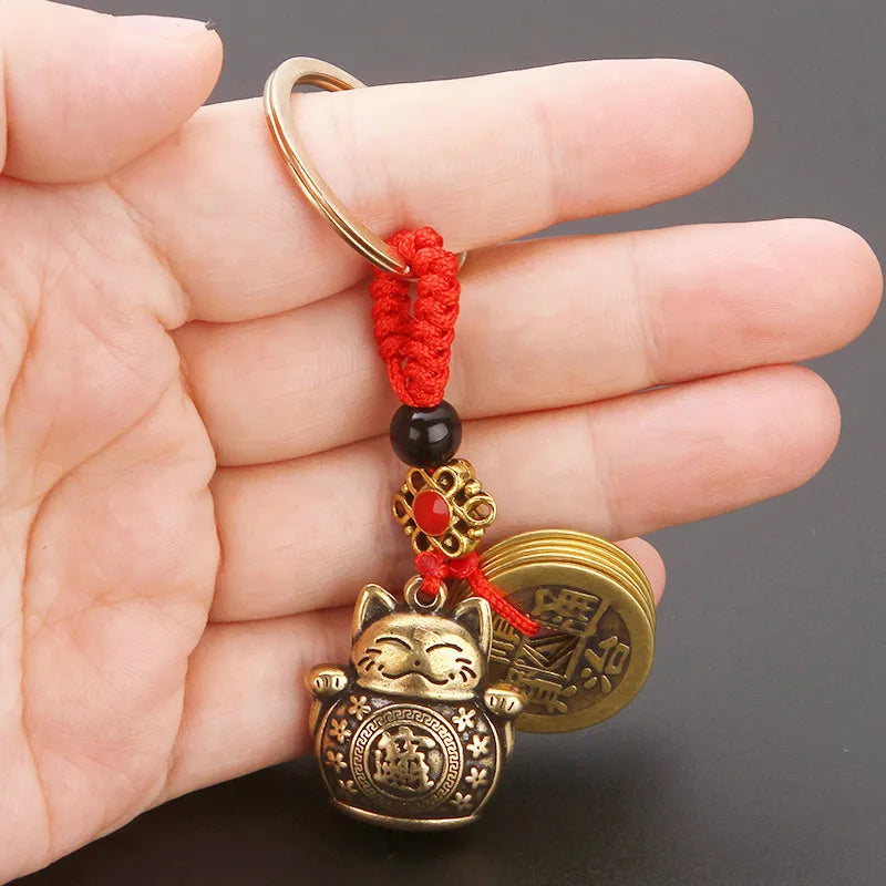 A vintage gold-tone lucky cat keychain with a red yarn tassel and gold coin charm held in a hand.