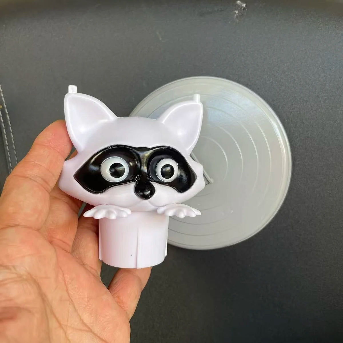 A white decorative toilet paper holder shaped like a cat's face with black markings, held in a hand against a gray background