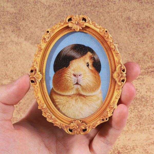 A hand holds an oval framed portrait of a brown and black lop-eared rabbit in an ornate gold frame.