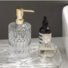 Klare Glasseife-Dispenserflasche mit goldfarbener Pumpe und eine kleine dunkle Flasche auf einer Glasplatte.