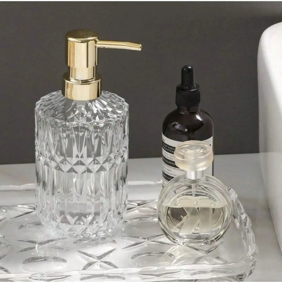 Klare Glasseife-Dispenserflasche mit goldfarbener Pumpe und eine kleine dunkle Flasche auf einer Glasplatte.