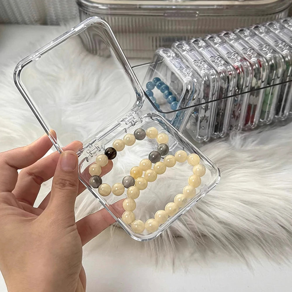 A hand holds a clear plastic jewelry organizer box filled with light yellow and gray beads, next to a clear plastic storage t