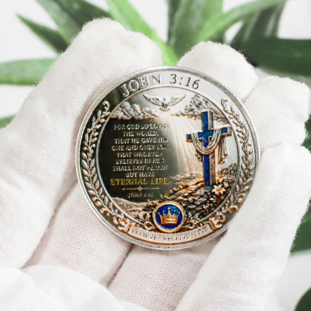 A hand holds a commemorative coin featuring John 3:16 and a cross design against a green leaf background.